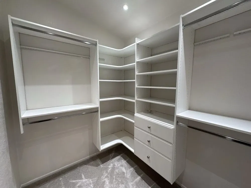 Custom white walk-in closet build-out with corner shelving, double hanging rods, and a drawer tower
